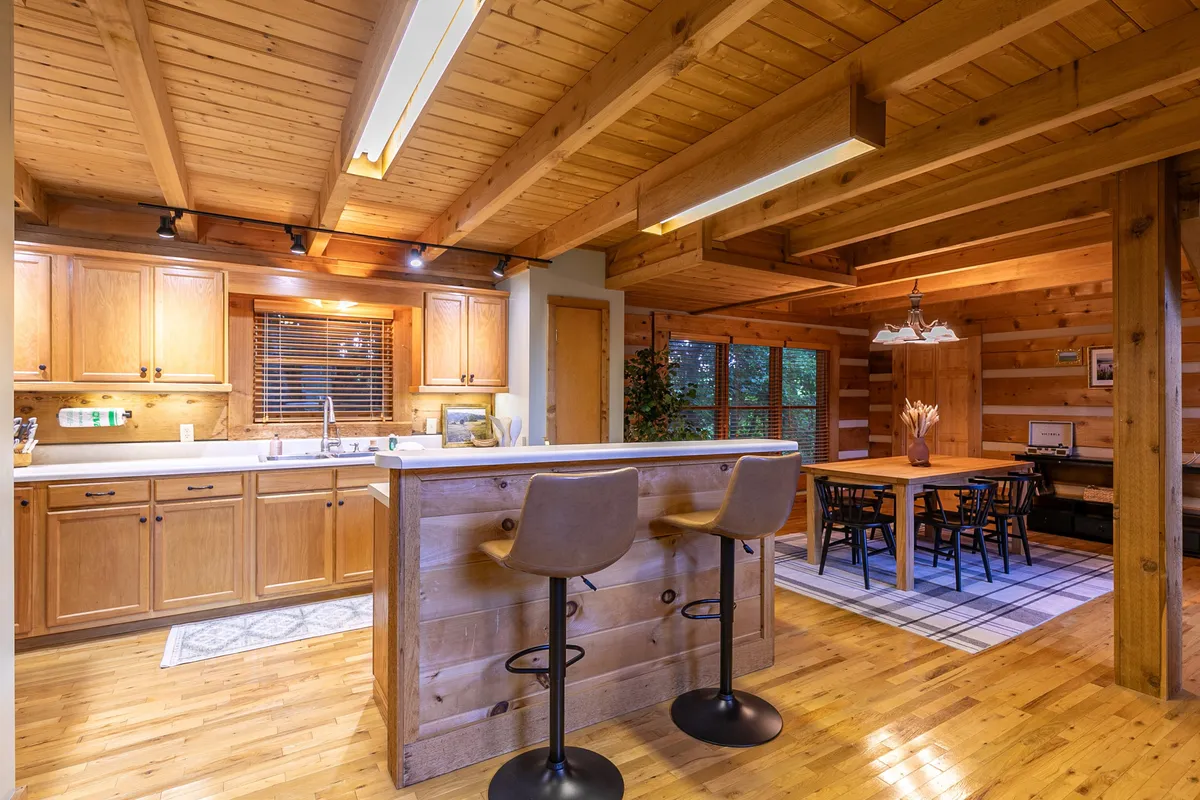A cozy, rustic kitchen and dining area with a kitchen island and bar stools, wooden cabinetry, a wooden dining table, and large windows with views of greenery. The space feels inviting and designed for both meals and socializing.