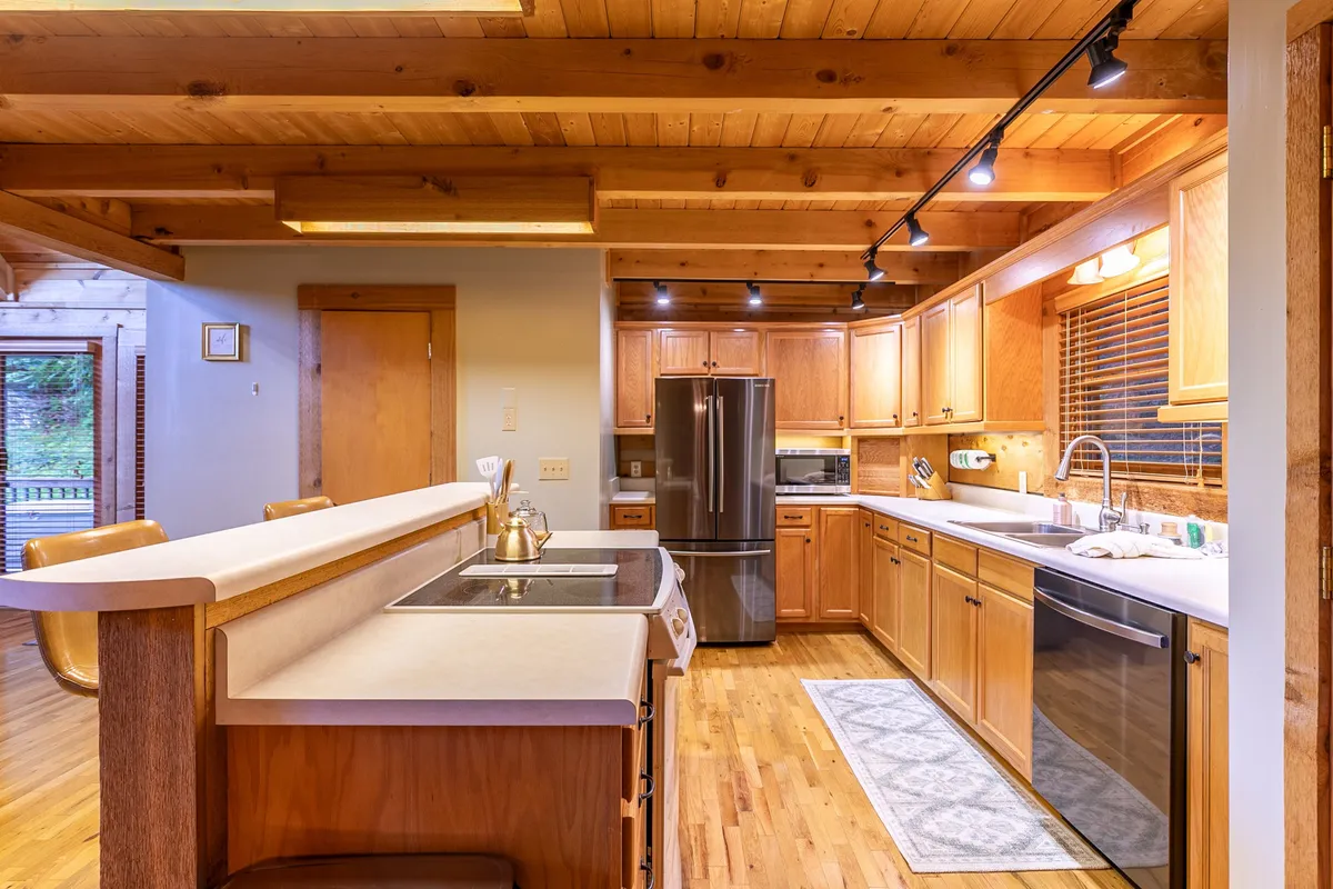 A bright, inviting kitchen with prominent wooden ceiling beams, a spacious kitchen island with barstools, and stainless steel appliances. Natural light floods in from the windows, illuminating the practical and stylish setup.