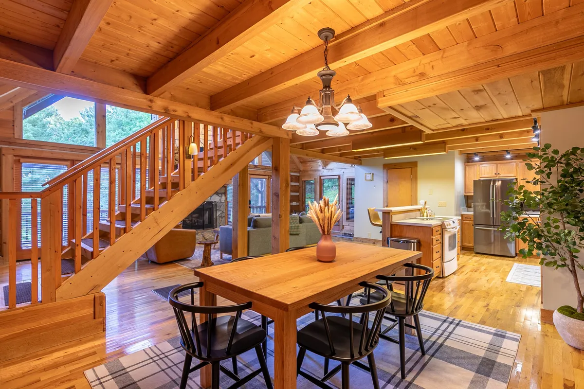 An open-plan space with a dining table adorned with a vase, a staircase leading upward, large windows framing forested views, and a fireplace for warmth. The space feels both communal and close to nature, ideal for socializing after outdoor adventures.