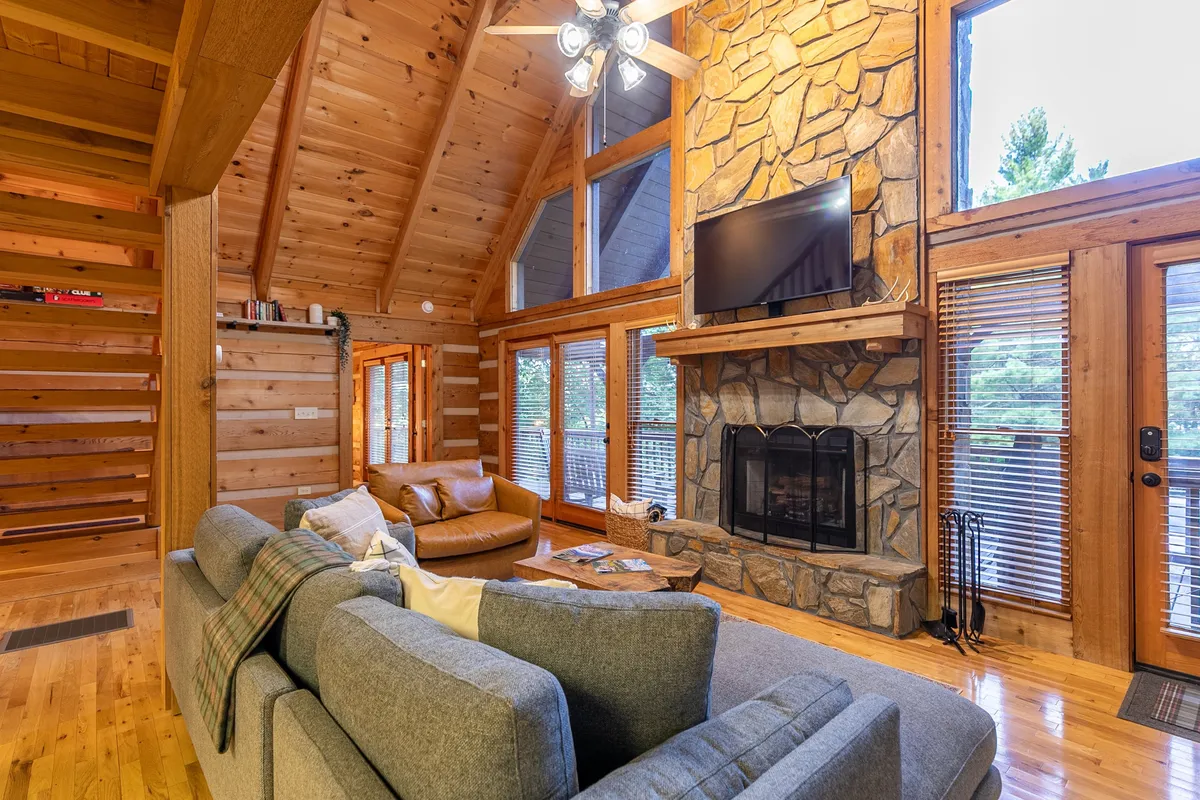 A spacious and comfortable seating area with a prominent stone fireplace, large windows that invite natural light and mountain vistas, and rustic wooden beams overhead. The layout encourages both socializing and relaxation.