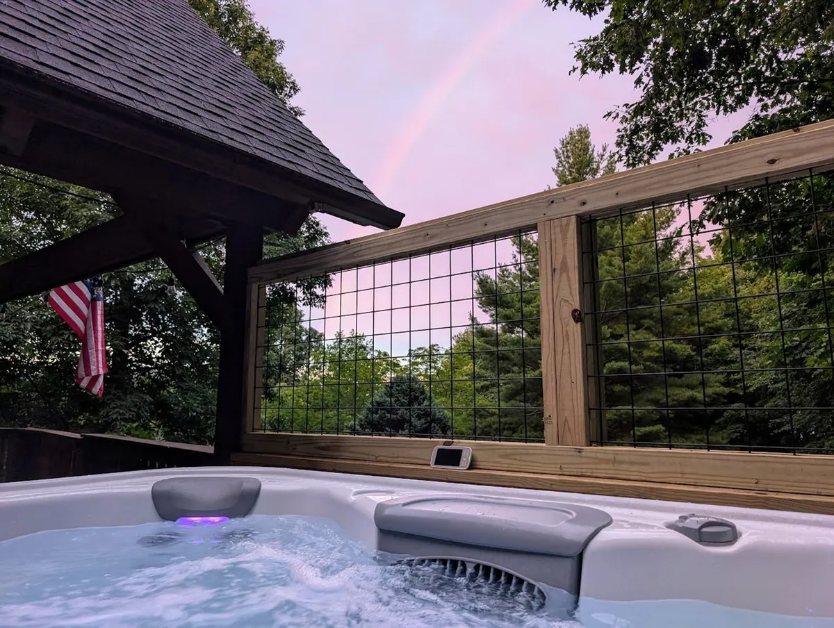 This image features the inviting hot tub set on the deck, surrounded by lush trees, with a rainbow arching in the background and an American flag fluttering. The tub is large enough for a group, and the serene atmosphere emphasizes relaxation and a connection with nature.