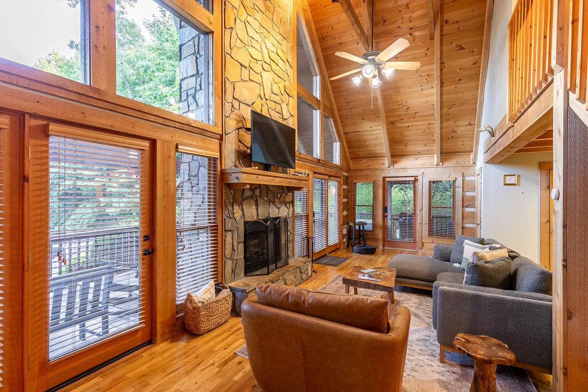 A spacious living area featuring a large stone fireplace, comfortable sofa seating, expansive windows revealing the lush mountain scenery, and prominent wooden beams overhead. The setting feels connected to the outdoors with natural light pouring in and a sense of rustic adventure.