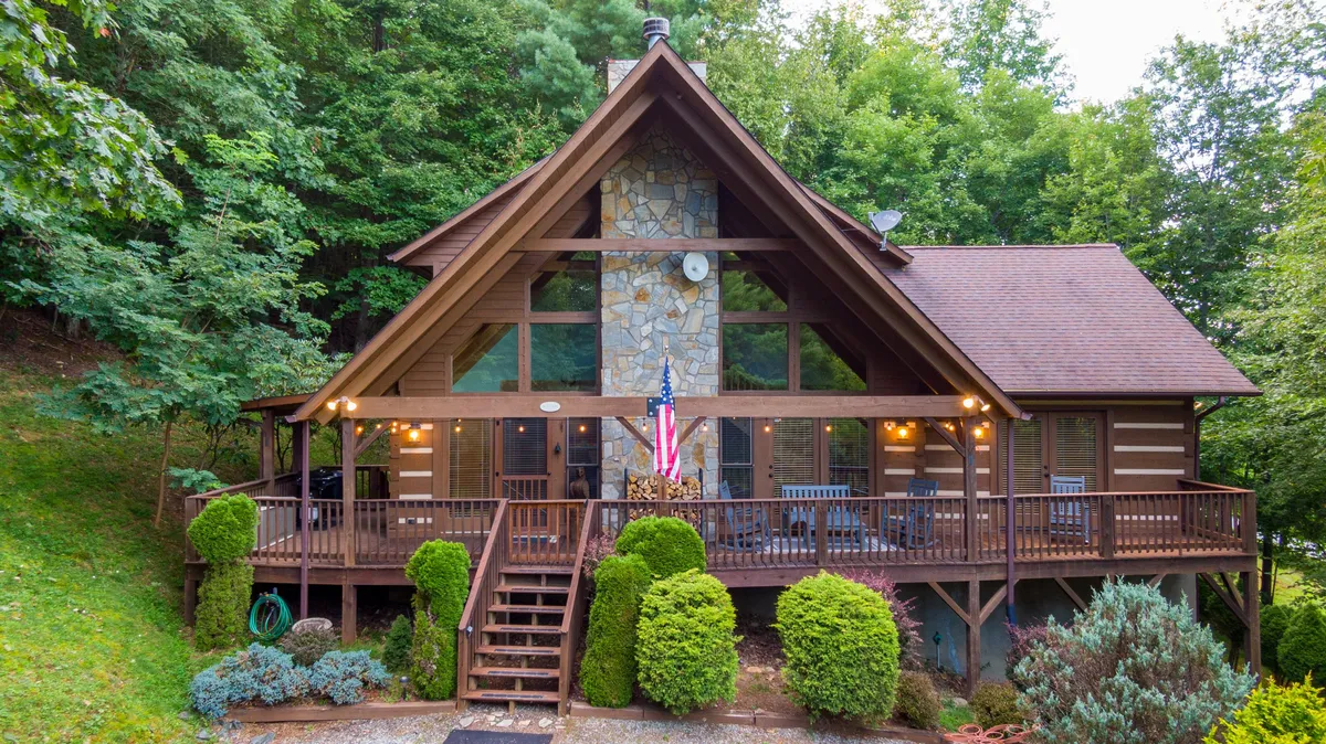The image shows a welcoming log cabin nestled in landscaped gardens, with a classic front porch perfect for relaxing. A stone chimney rises on one side, while an American flag flutters nearby, evoking a sense of tradition and warmth. The setting is tranquil, surrounded by the natural beauty of the North Carolina mountains.