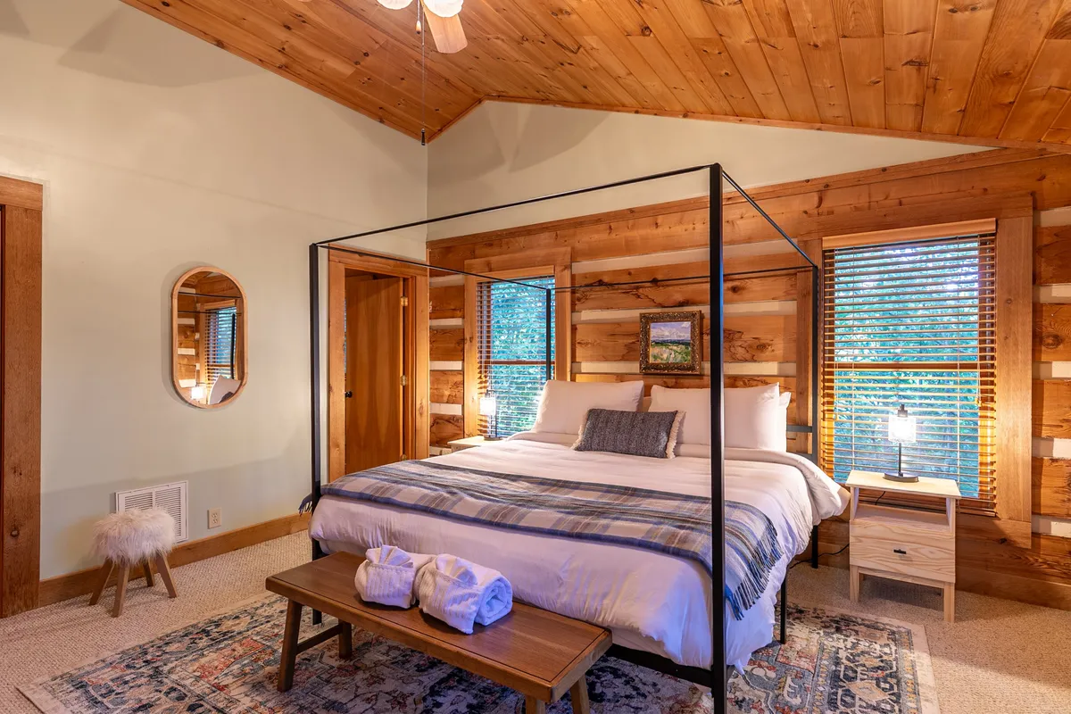 A cozy bedroom with a canopy bed framed by warm wooden walls, a nightstand with a lamp, and a window letting in gentle natural light. The atmosphere is tranquil and inviting, perfect for relaxing after a day outdoors.