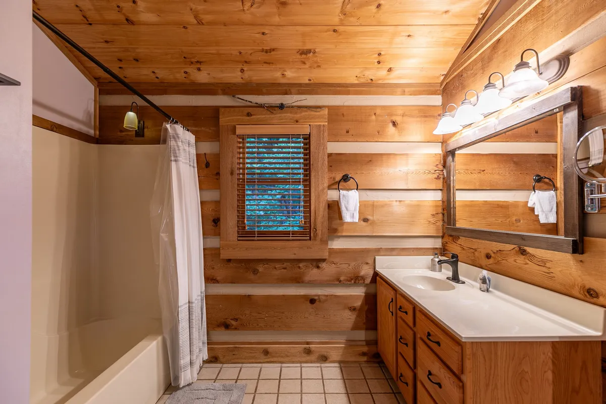 A cozy bathroom with wooden walls and a window fitted with blinds, allowing for natural light to enter. The sink area and shower look inviting—ideal for refreshing after a day spent outdoors.