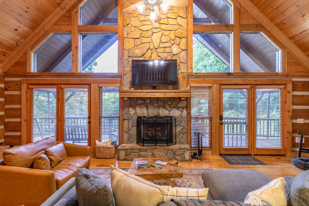 A welcoming great room with a large stone fireplace, expansive windows revealing glimpses of mountain scenery, and cozy seating arranged for conversation or relaxation. The space feels open and bright, suggesting a strong connection to the outdoors.