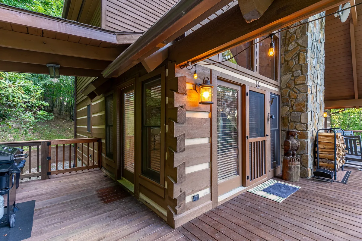 The deck area at dusk illuminated by soft outdoor lighting, with visible wooden logs, a stone wall, and seating. This welcoming setup hints at the potential for grilling, socializing, and enjoying the ambiance as the sun sets over the mountains.