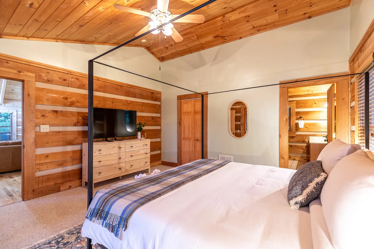 An elegant four-poster bed is the focal point in this tranquil room, complemented by a wooden dresser, decorative mirror, and a flat-screen TV. Soft lighting and tasteful decor enhance the relaxing ambiance.