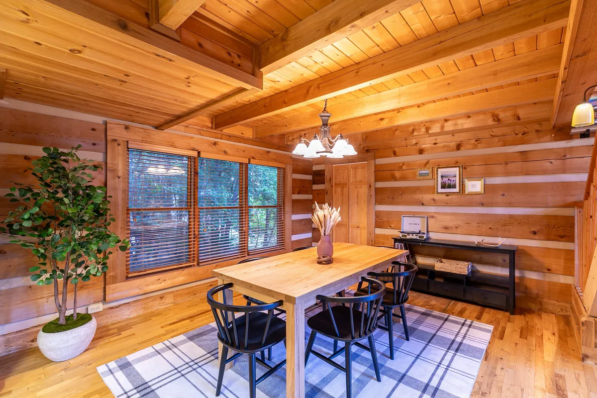 A warmly lit dining area with a sturdy wooden table set beneath a rustic chandelier. Large windows dominate the scene, allowing ample natural light to pour in and showcasing glimpses of the outdoors. An indoor plant adds a touch of greenery, and the space feels both welcoming and connected to nature.