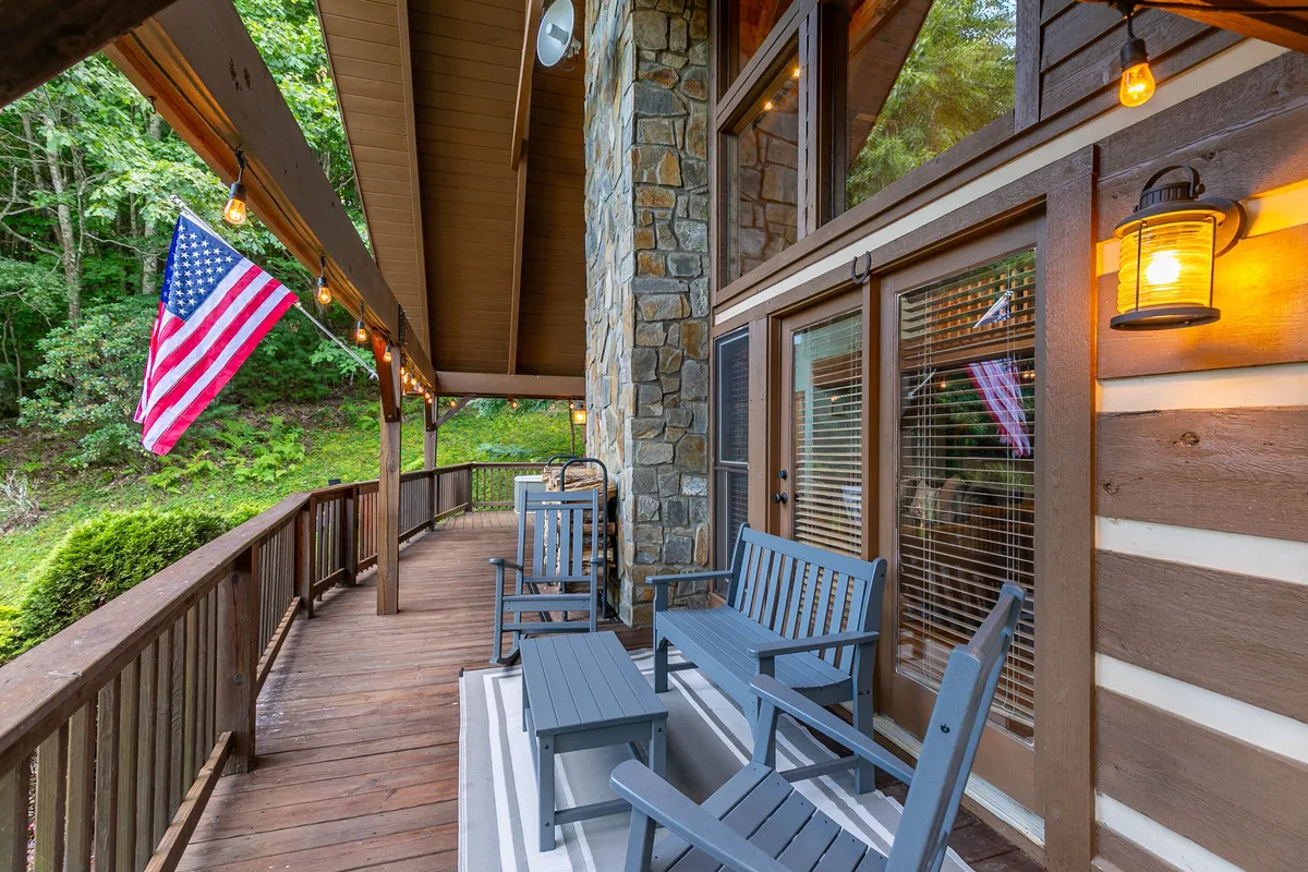 The image shows a welcoming porch area with American flag, stone wall, and wooden rocking chairs, all illuminated by porch lighting. The main entry is implied just behind, suggesting the home’s accessible entryway.