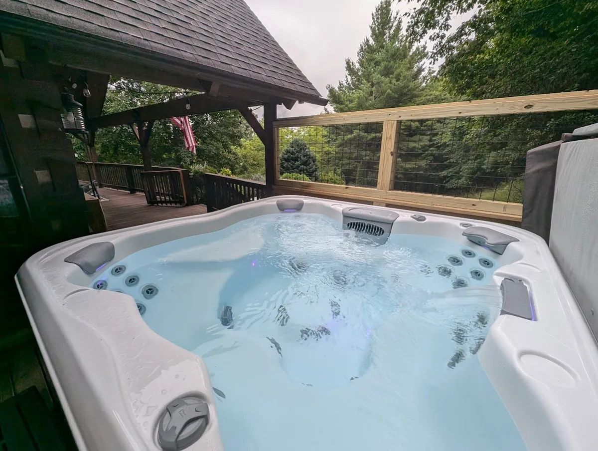 A tranquil scene of the wooden deck featuring the hot tub set amidst surrounding greenery. The deck area is expansive, with ample space for seating and enjoying the fresh mountain air. The atmosphere is peaceful and perfect for morning coffee or evening relaxation.