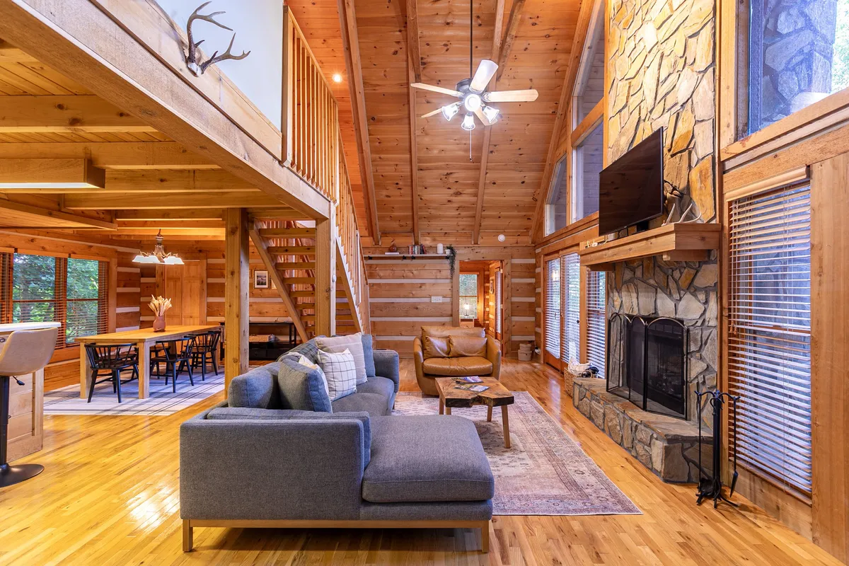 A warm, open great room featuring a large stone fireplace as a focal point. Comfortable sofa seating is arranged to encourage conversation and relaxation, with large windows letting in natural light. The wooden beams overhead add rustic charm. A dining table for six is visible, seamlessly connecting the living and dining spaces.